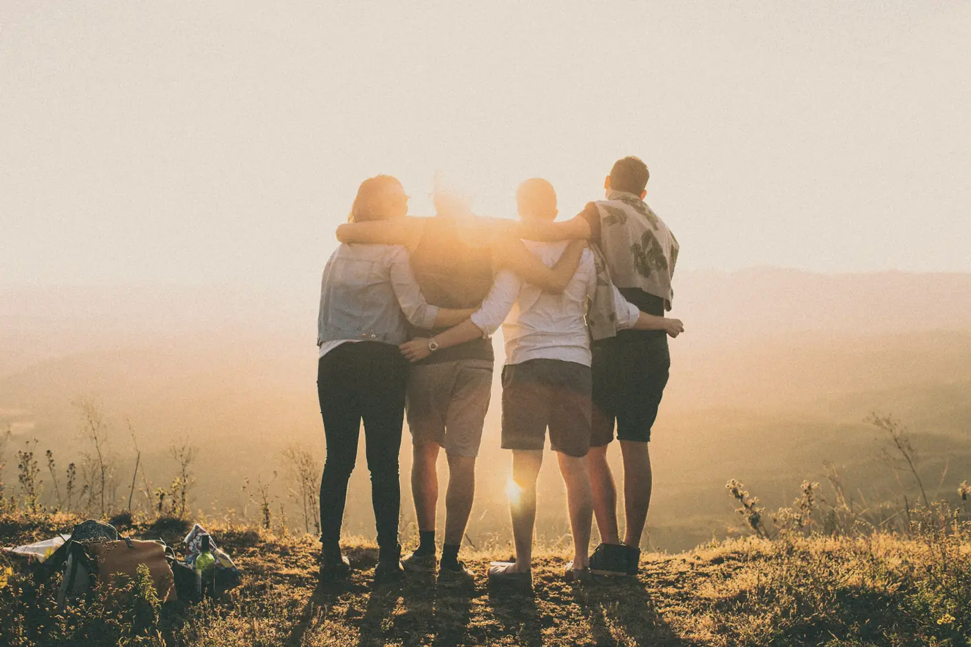 Four friends with their arms around each other, watching the sun set over the hills